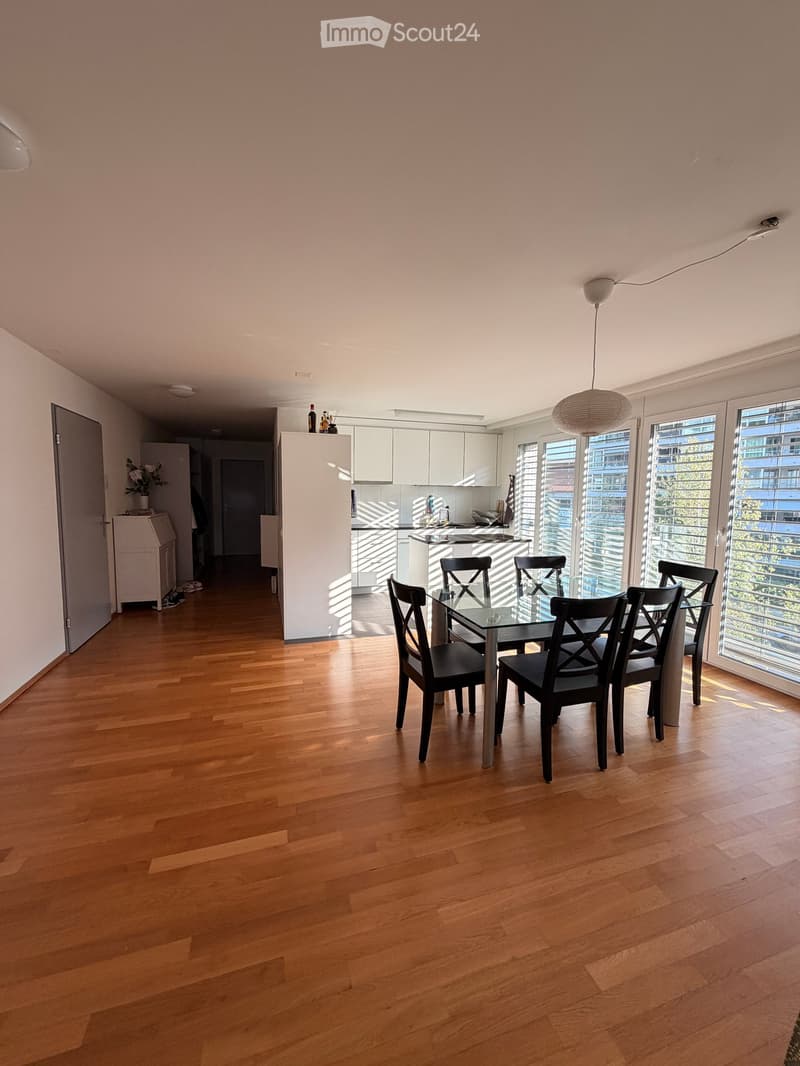 Dining area with a glass dining table and black chairs, adjacent to a modern kitchen with white cabinets and stainless steel appliances, large windows with blinds, and wooden flooring.