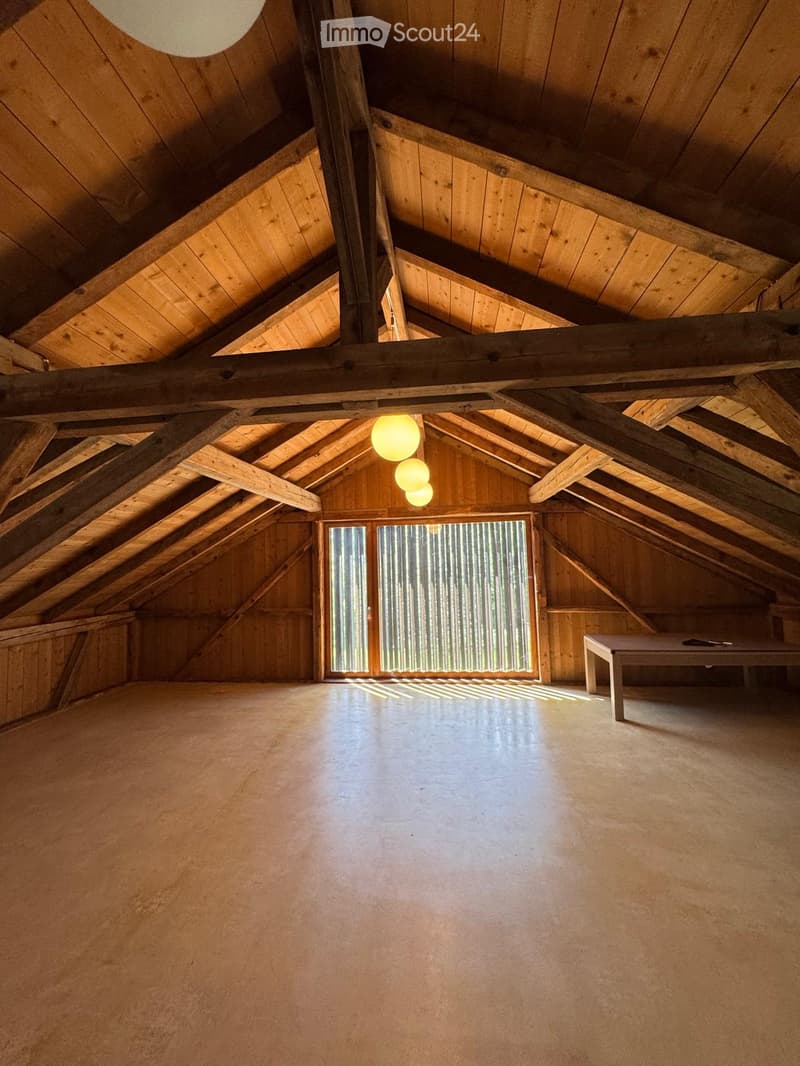 An attic space with exposed wooden beams and a high, sloped wooden ceiling. The room features a large window with vertical blinds, concrete flooring, and a single small table with a few items on it.