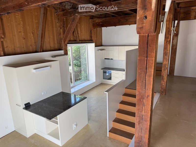 Open-plan kitchen and living area with white cabinets, dark countertops, and wooden beams. Features a staircase leading to an upper level.