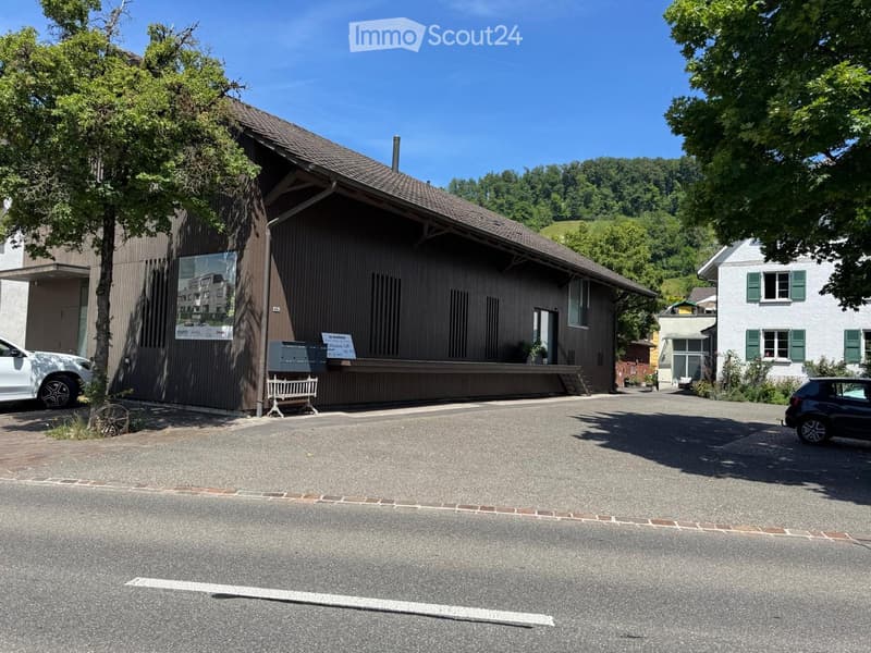 A large wooden building with a sloped roof, surrounded by trees and parked cars. The building has a signboard and a small bench near the entrance.