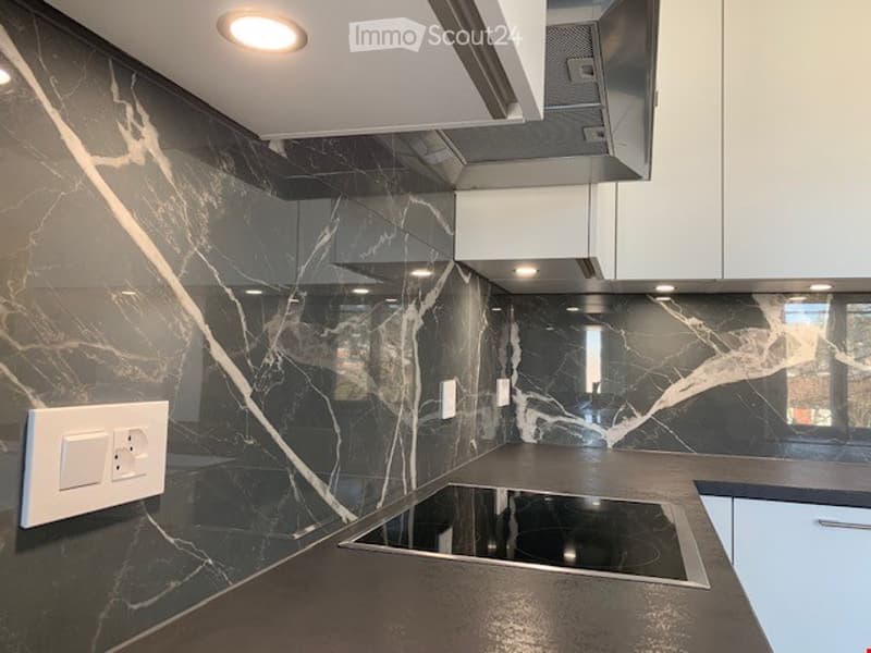 Modern kitchen with dark marble backsplash, sleek black countertops, and built-in induction cooktop. White cabinets and stainless steel range hood are visible.