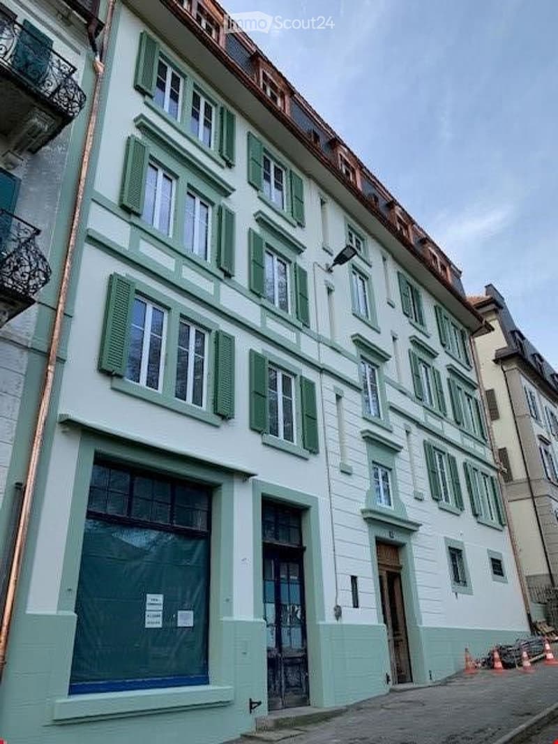 Four-story residential building with white facade, green shutters, and multiple windows. The ground floor has a covered entrance and a secured window with protective bars.