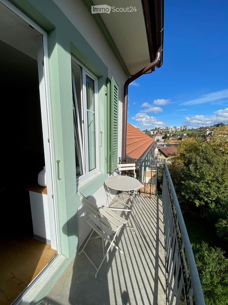Balcony with a round table and two chairs, overlooking a cityscape with trees and buildings.