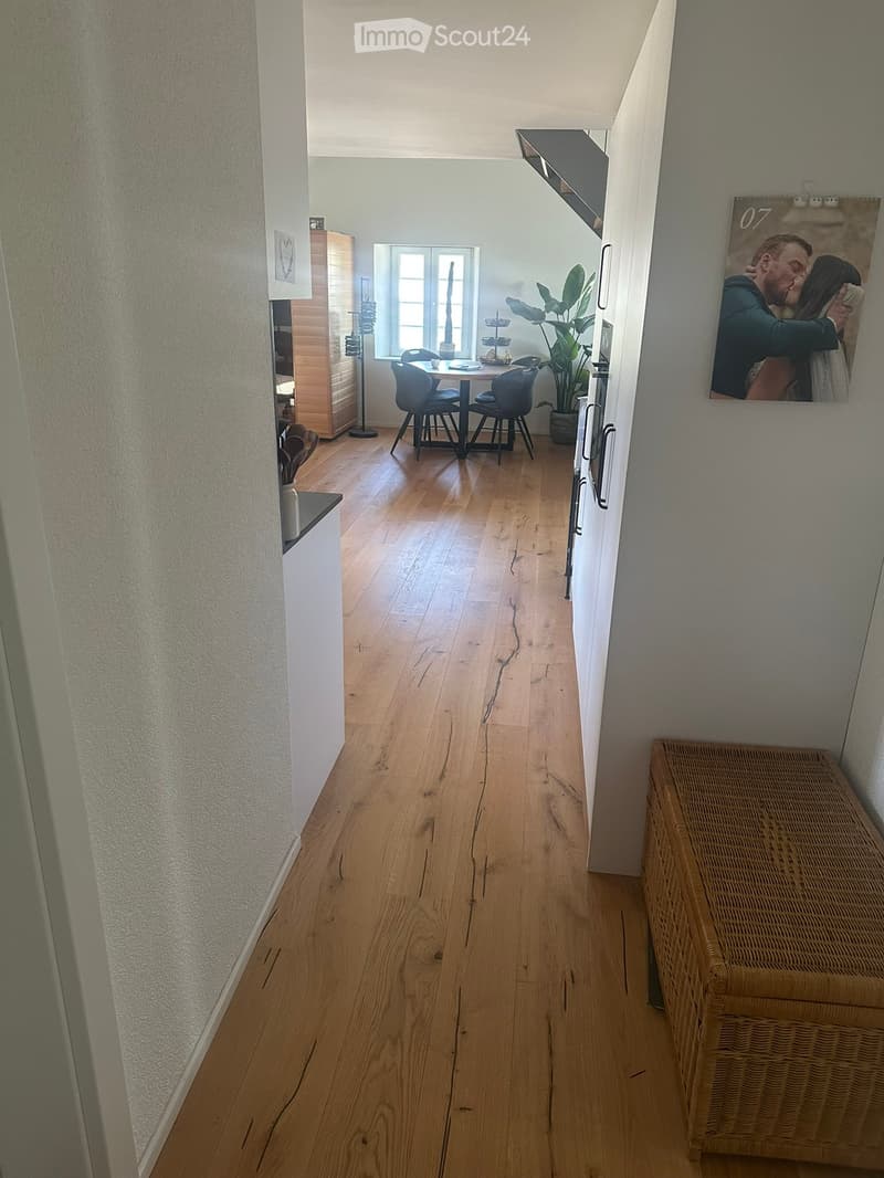 A narrow hallway with wooden flooring, white walls, and a wicker bench on the right side. A doorway leads to a dining area with a table, chairs, and a potted plant.