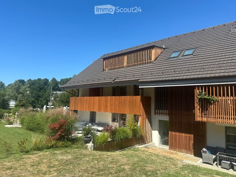 Modern two-story house with wooden balconies, large windows, and a well-maintained garden with seating area.