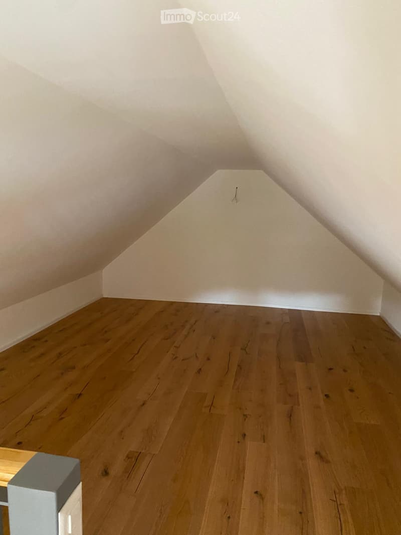 Empty attic space with wooden flooring and slanted white ceilings.