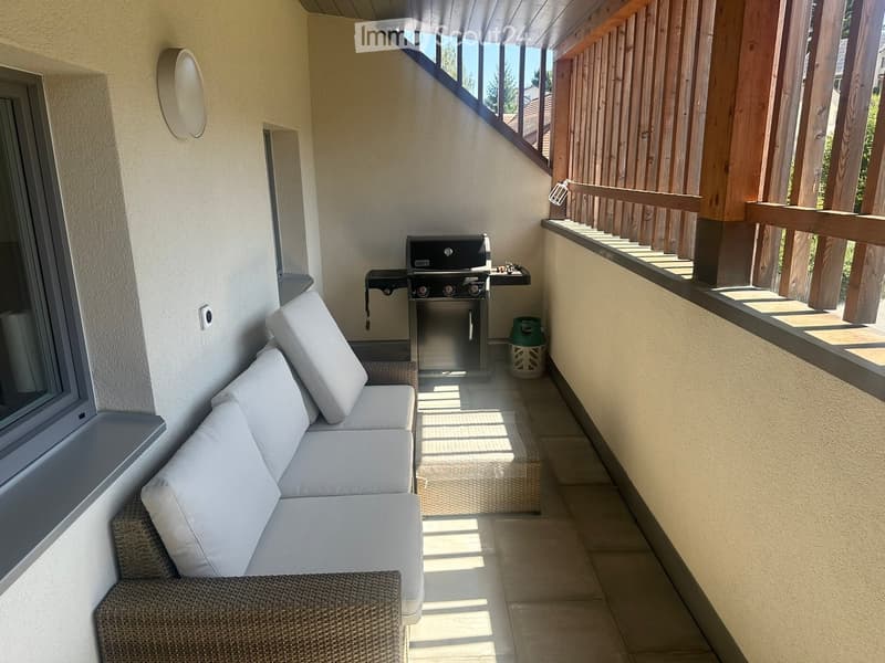 Balcony with a white sofa, a barbecue, and a small wicker stool. The balcony has wooden railings and tiled flooring.