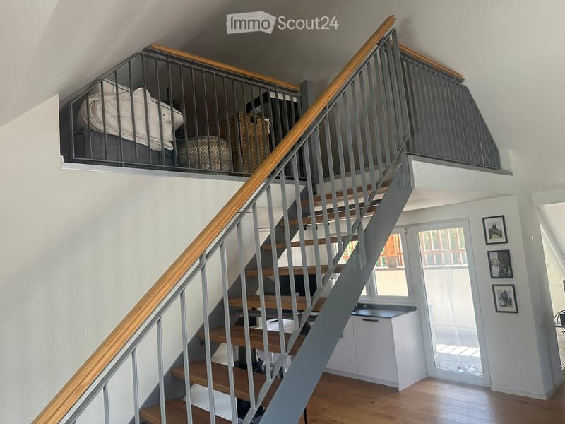 Staircase with wooden handrail and metal balustrade, leading to a loft area with a bed and storage baskets. The hallway features white walls, wooden flooring, and framed pictures on the wall.
