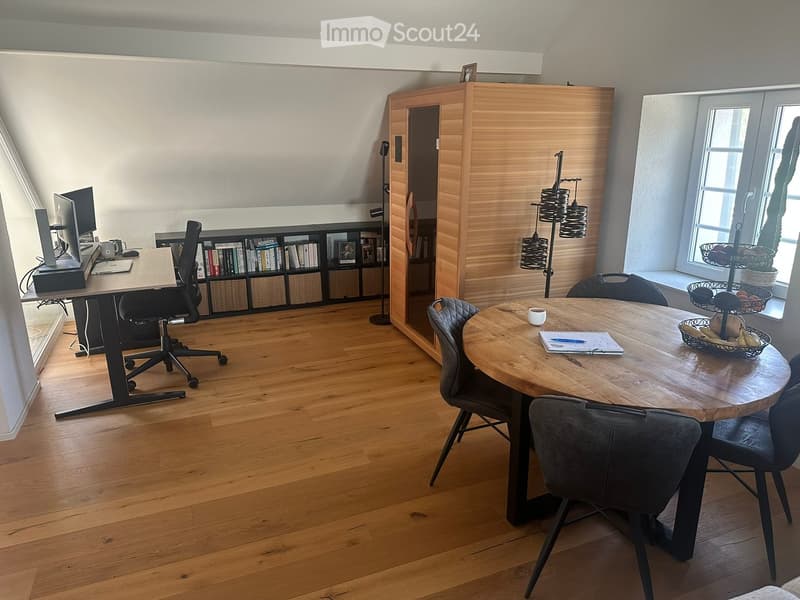 A modern, well-lit room with a wooden floor, featuring a desk with a laptop and chair, a round wooden table with chairs, a bookshelf, and a floor lamp.
