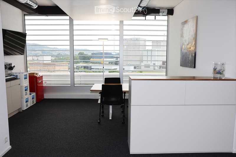Modern office space with large windows, a reception desk, a chair, a table, a refrigerator, and a screen.