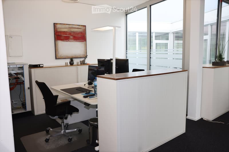 Modern office space with a white desk, black office chair, computer setup, and a reception area with a white counter. Large windows provide natural light, and there is a small plant and artwork on the wall.
