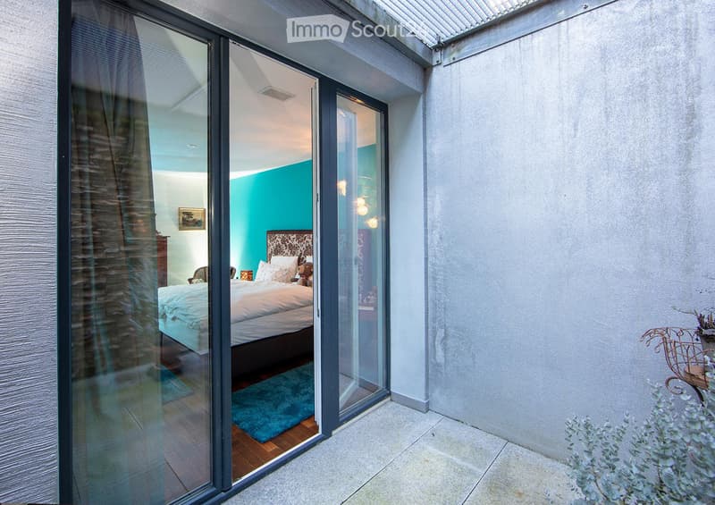 Modern bedroom with large glass sliding doors opening to a balcony, featuring a bed, nightstand, chair, and picture frame on the wall.