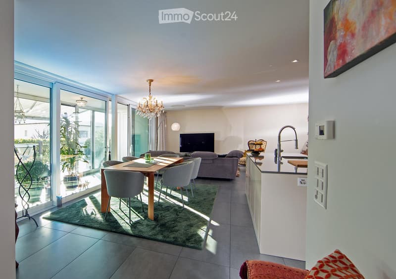 Open-plan living and dining area with large glass doors leading to a balcony, a sofa, dining table with chairs, television, chandelier, floor lamp, and a modern kitchen area with a faucet.