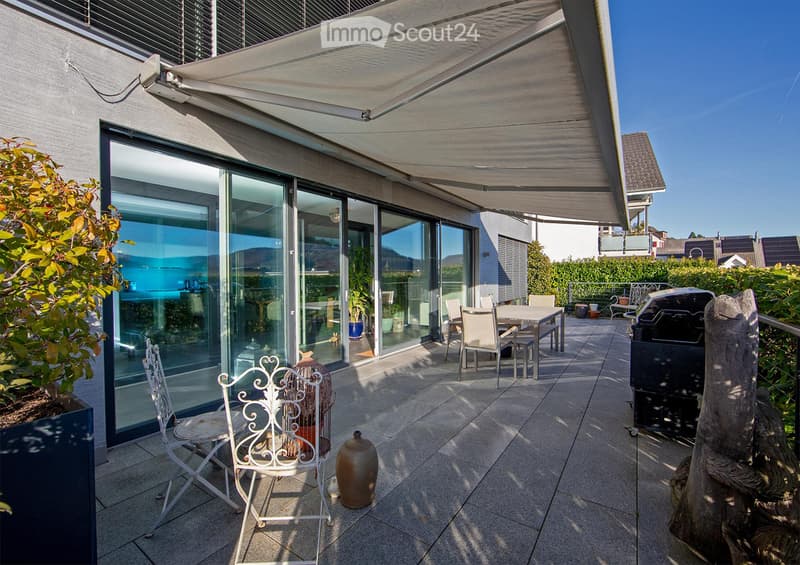 A modern, covered balcony with large glass sliding doors leading to an indoor space. The balcony features a dining table with chairs, a grill, and decorative elements like vases and a wooden sculpture.