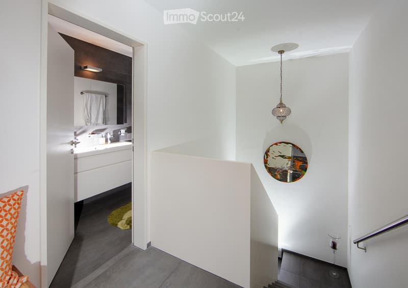 Modern hallway with white walls, grey tiled floor, a decorative round mirror, a hanging light fixture, and an open door leading to a bathroom with white cabinets and a sink.