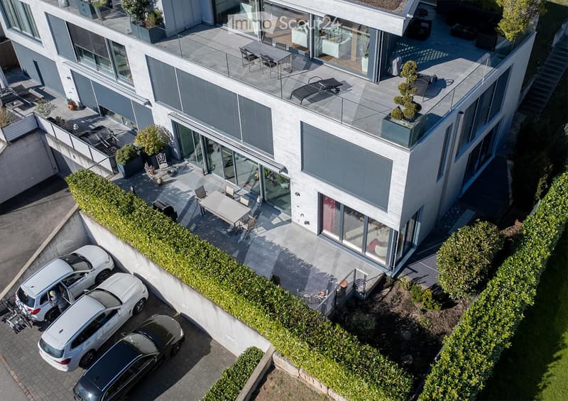Modern multi-story building with large glass windows, balconies, and outdoor seating areas. Includes a parking area with several cars.