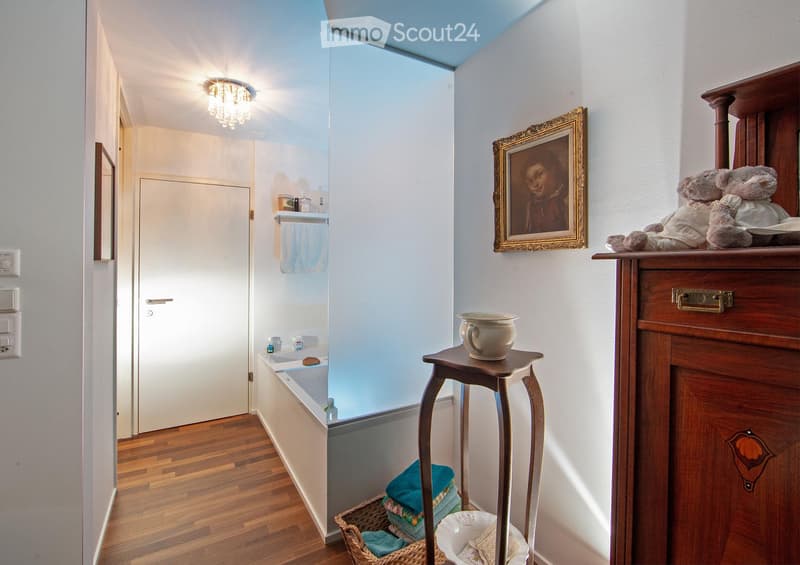 Bathroom with wooden floor, bathtub, small stool with cup, wooden cabinet with framed picture and teddy bears, wicker basket with towels.