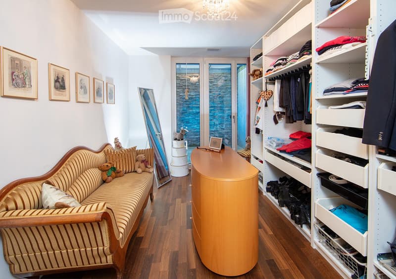 A cozy, well-furnished room with a striped sofa, decorative pillows, a large mirror, a modern wardrobe with shelves and drawers, and framed artwork on the walls.