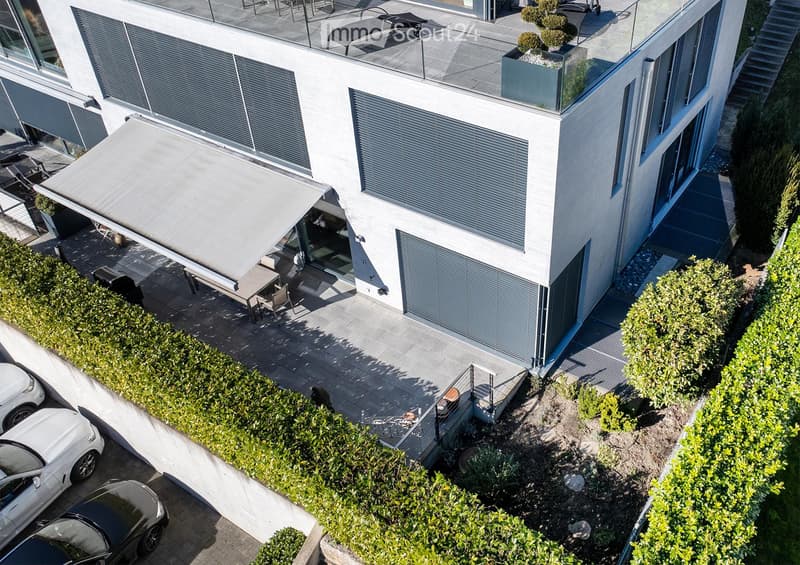 Modern two-story building with a rooftop terrace, outdoor seating, and a covered patio area. The building features large windows and a sleek design. There is a parking area with several cars visible.