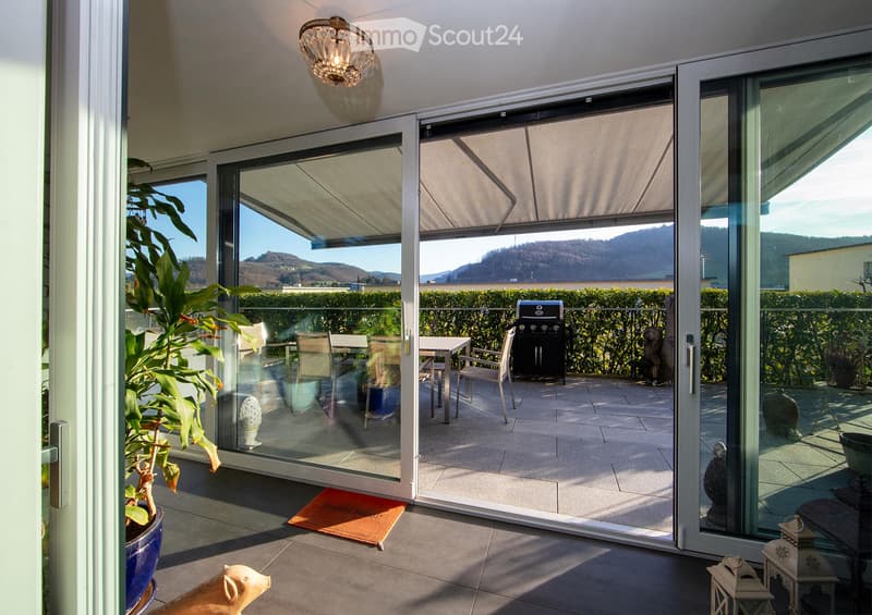 Spacious balcony with large glass sliding doors, outdoor dining set, grill, and scenic mountain view.