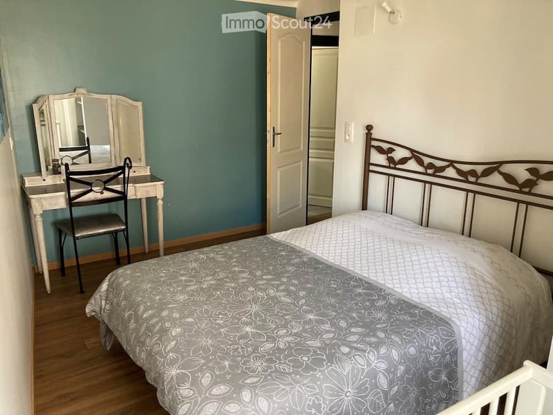 Cozy bedroom with a wooden bed, floral bedspread, vanity with mirror, and a chair. The room features teal walls and wooden flooring.