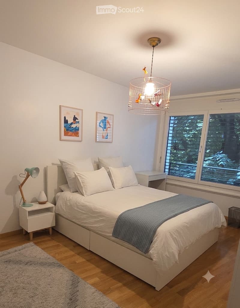 Modern bedroom with a large bed, white bedding, nightstand with a lamp, and a chandelier hanging from the ceiling. The room features large windows with blinds and a view of greenery outside.