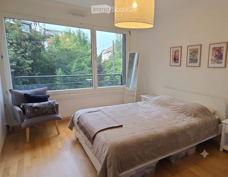 Bright bedroom with wooden flooring, large windows offering a view of greenery, a double bed with a beige duvet, a grey armchair with patterned cushions, a mirror, and three framed artworks on the wall.
