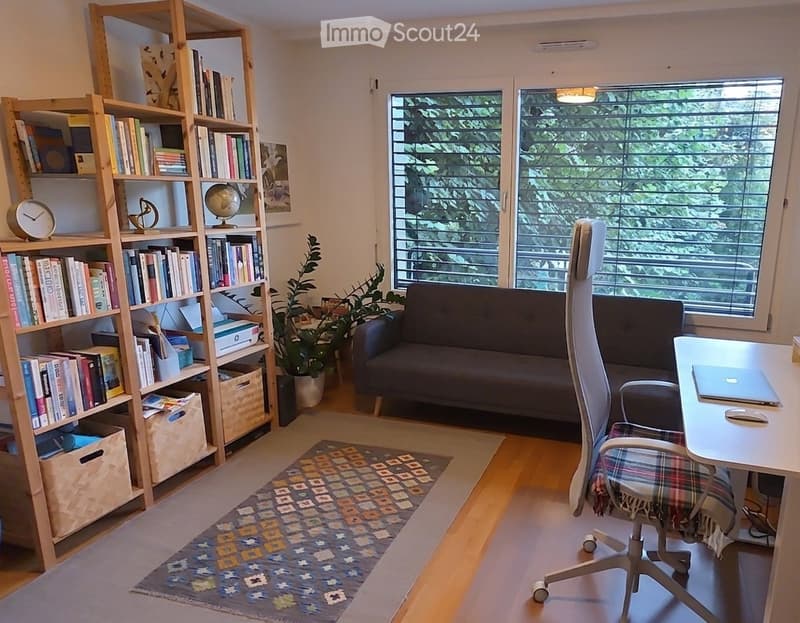Cozy living room with a dark grey sofa, a white desk with a laptop, a wooden bookshelf filled with books, a globe, a clock, and a potted plant near the window offering a view of greenery.