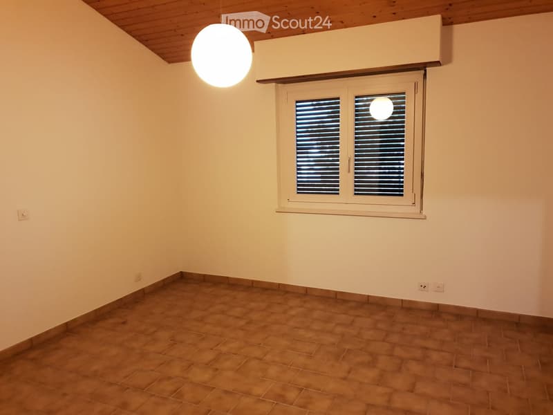 Empty room with beige tiled floor, white walls, and a single window with shutters. A round pendant light hangs from the ceiling.