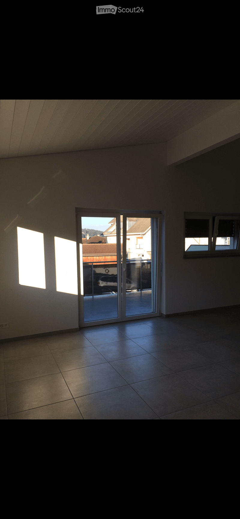 Empty room with tiled floor, large sliding glass door leading to a balcony, and windows allowing natural light.