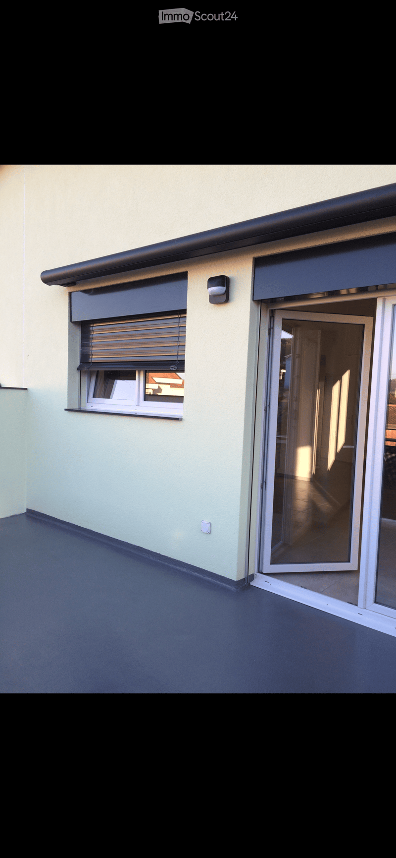 Balcony with a dark grey floor, light green wall, sliding glass door, window with roller blinds, and a small light fixture.