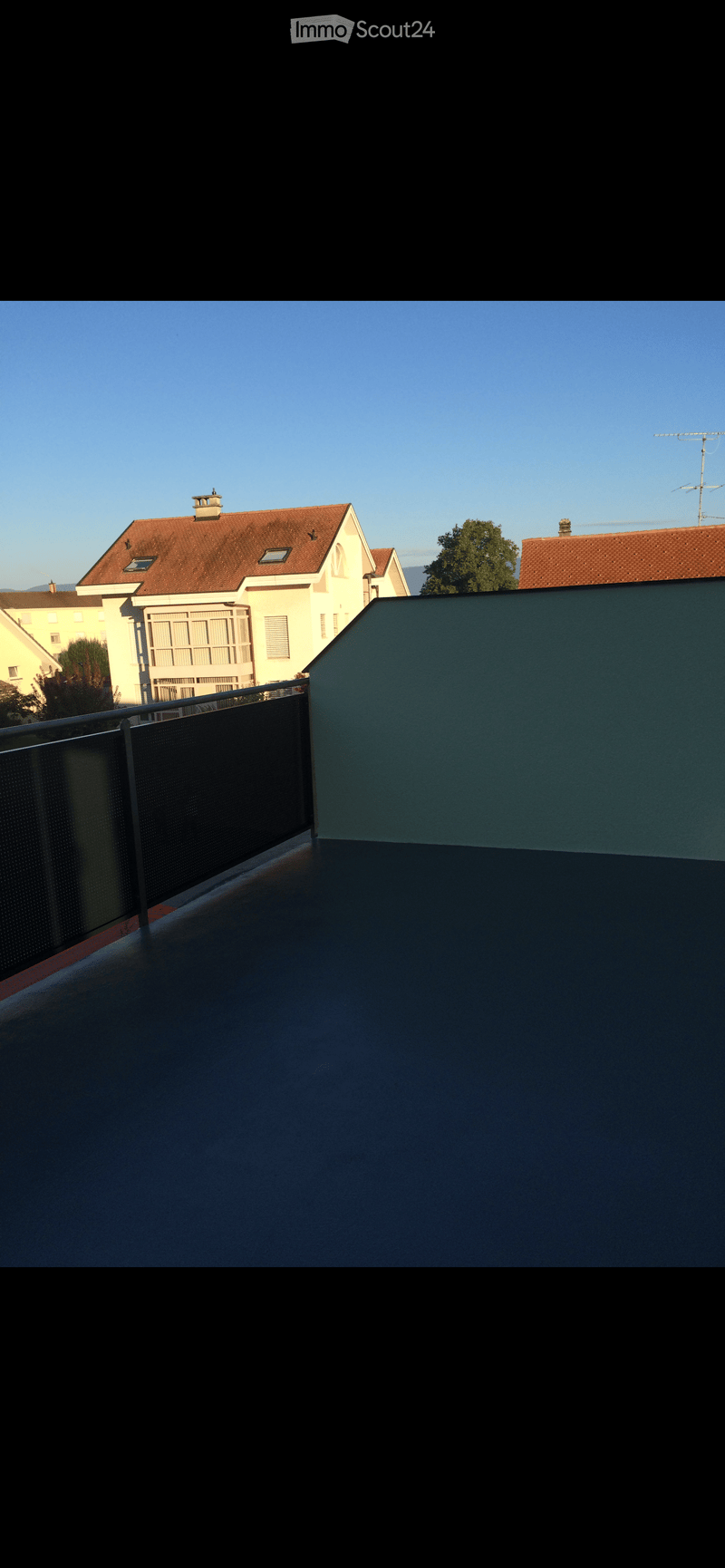 Roof terrace with a metal railing, overlooking neighboring houses with red-tiled roofs and a clear blue sky.