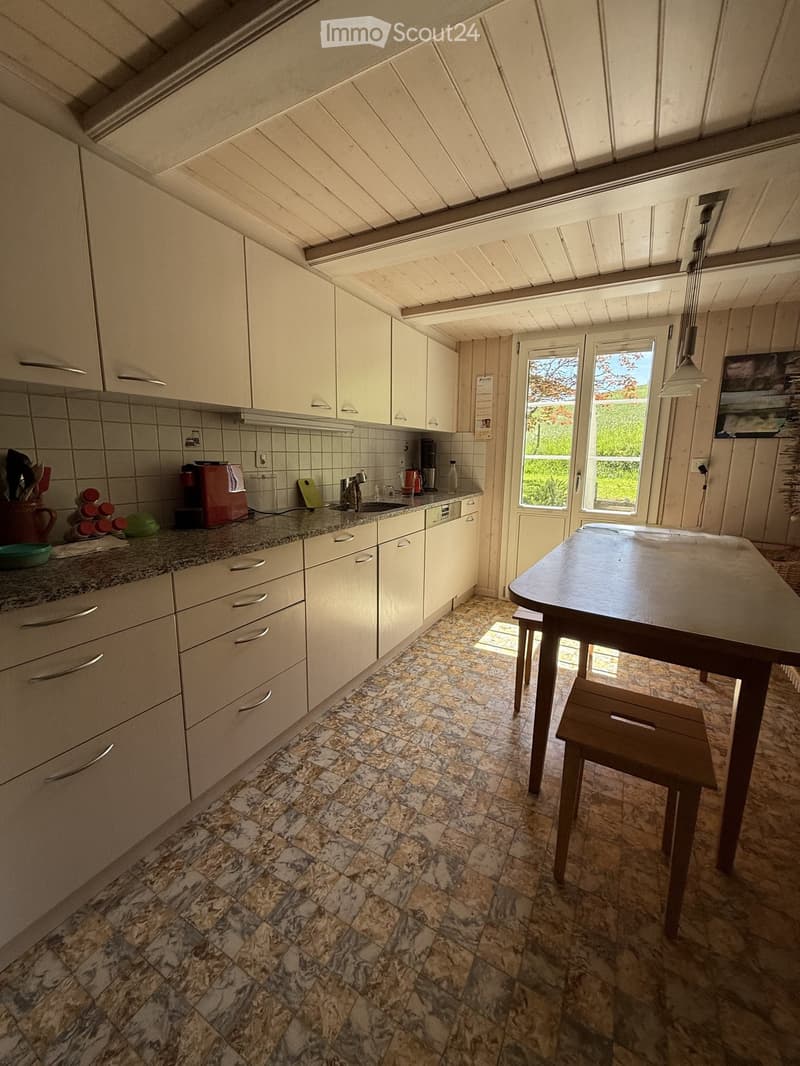 Bright kitchen with white cabinets, granite countertops, and a wooden dining table with a single stool. The space features a sink, coffee maker, and various kitchen items on the counter.