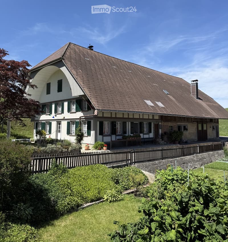 Two-story house with a pitched roof, white walls, green shutters, and a covered porch area with outdoor seating and potted plants.