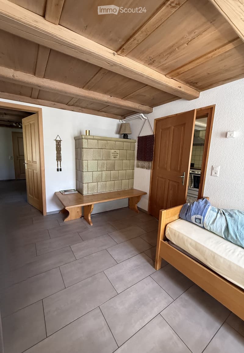 Cozy attic bedroom with wooden slanted ceilings, tiled floor, a wooden bed with bedding, a wooden bench, a lamp, and a mirror. A door leads to another room.