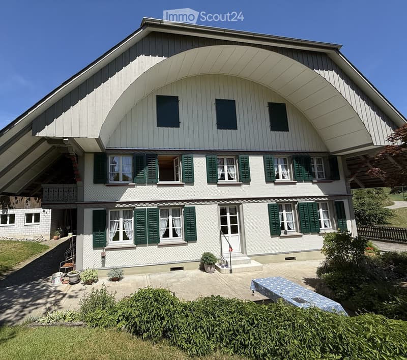 Two-story house with white siding, green shutters, and multiple windows. Features a raised ground floor, a balcony, and a well-maintained garden area with potted plants and a covered patio.