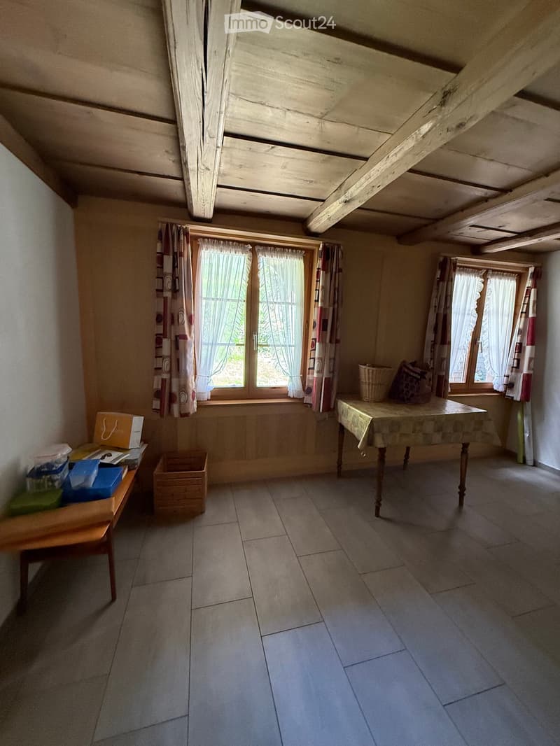Room with exposed wooden beams, tiled floor, windows with curtains, a small table with a cloth, a chair, and a basket on the windowsill.