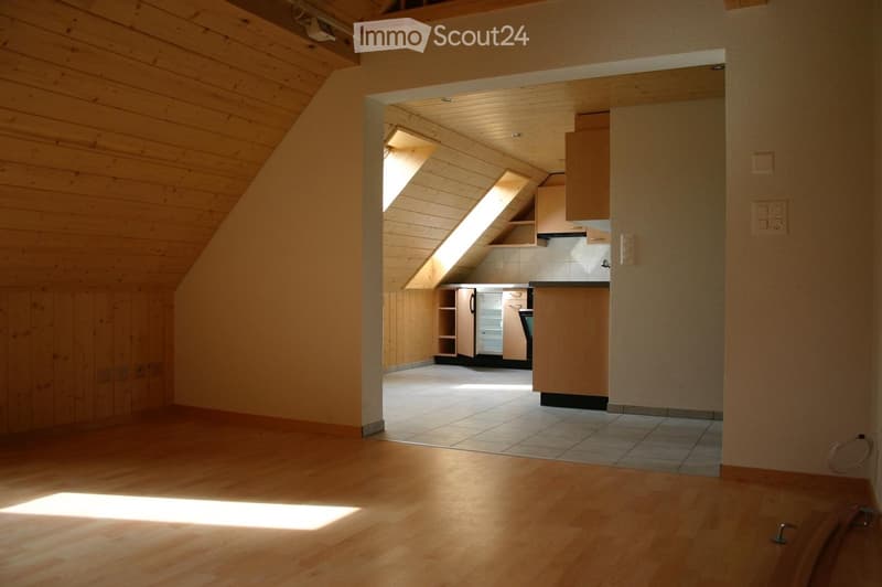 Bright, wooden-floored room with exposed wooden beams, leading to a small kitchen area with cabinets, a refrigerator, and a sink.