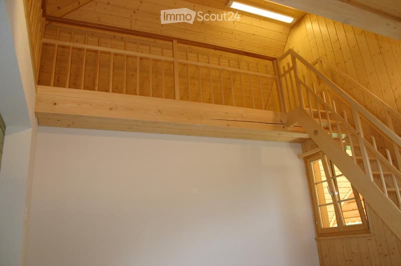 Wooden staircase leading to an upper level with a wooden railing and exposed wooden beams on the ceiling. The walls are white, and there is a small window at the end of the hallway.