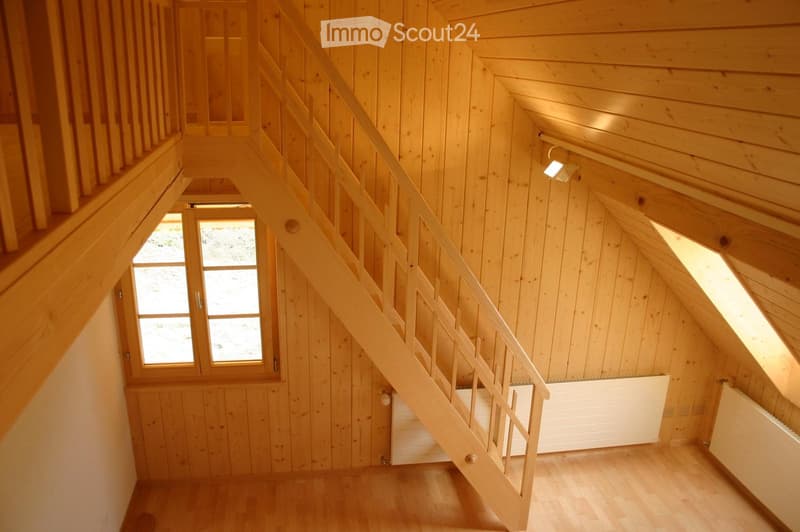 Wooden staircase with a handrail, wooden walls, and a skylight window providing natural light. The floor is made of light-colored wood, and there are radiators on the walls.