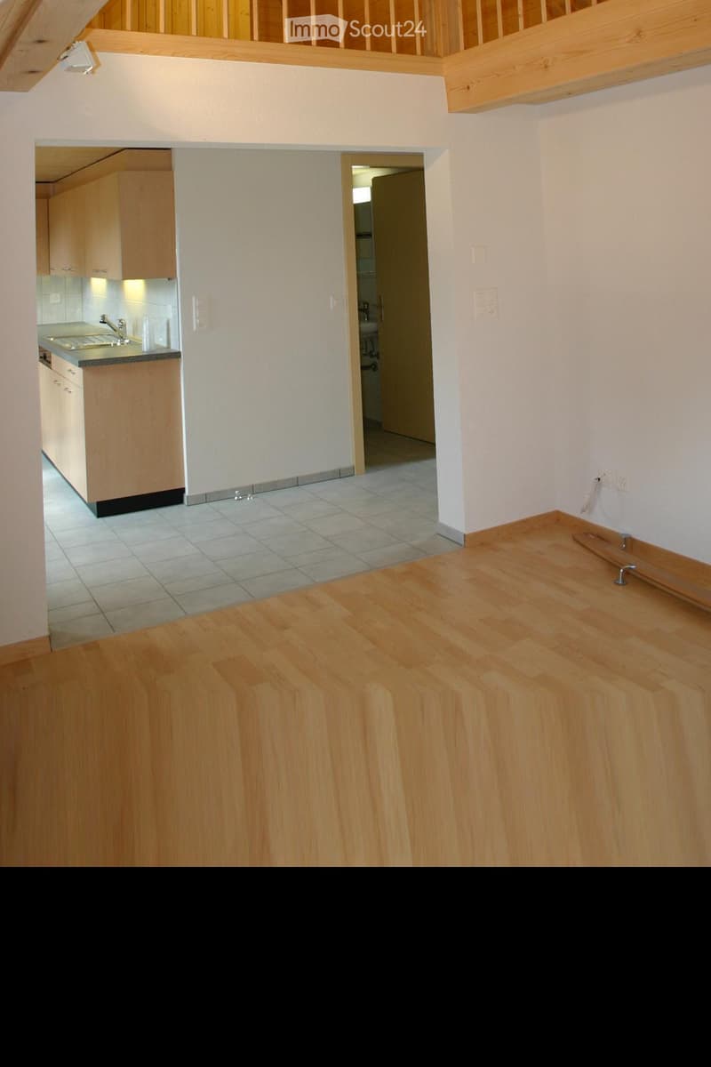 Empty room with wooden flooring, white walls, and a small kitchen area visible in the background. The kitchen has light wooden cabinets and a sink.