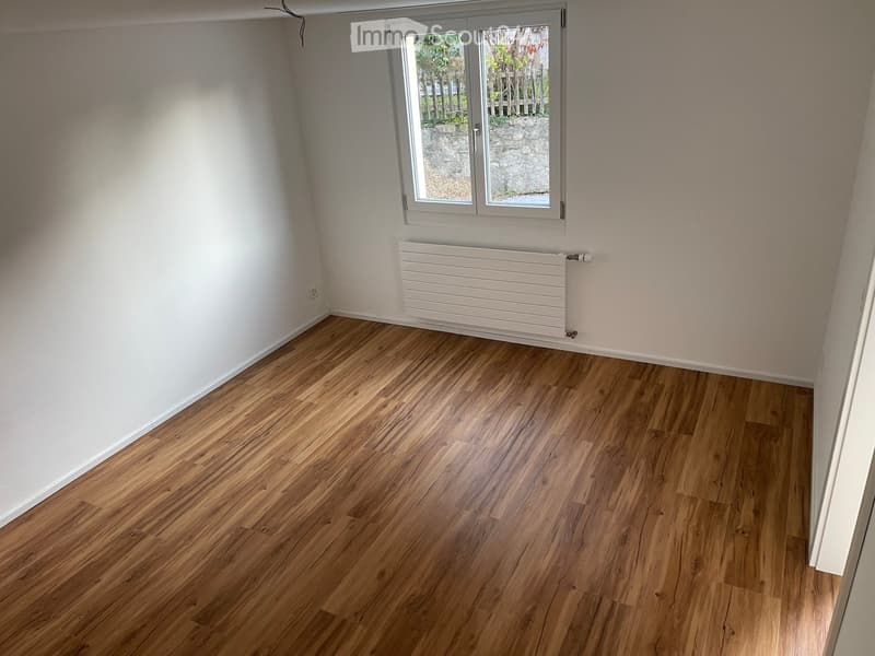 Empty room with wooden flooring, white walls, a window with a view of a garden, and a white radiator below the window.