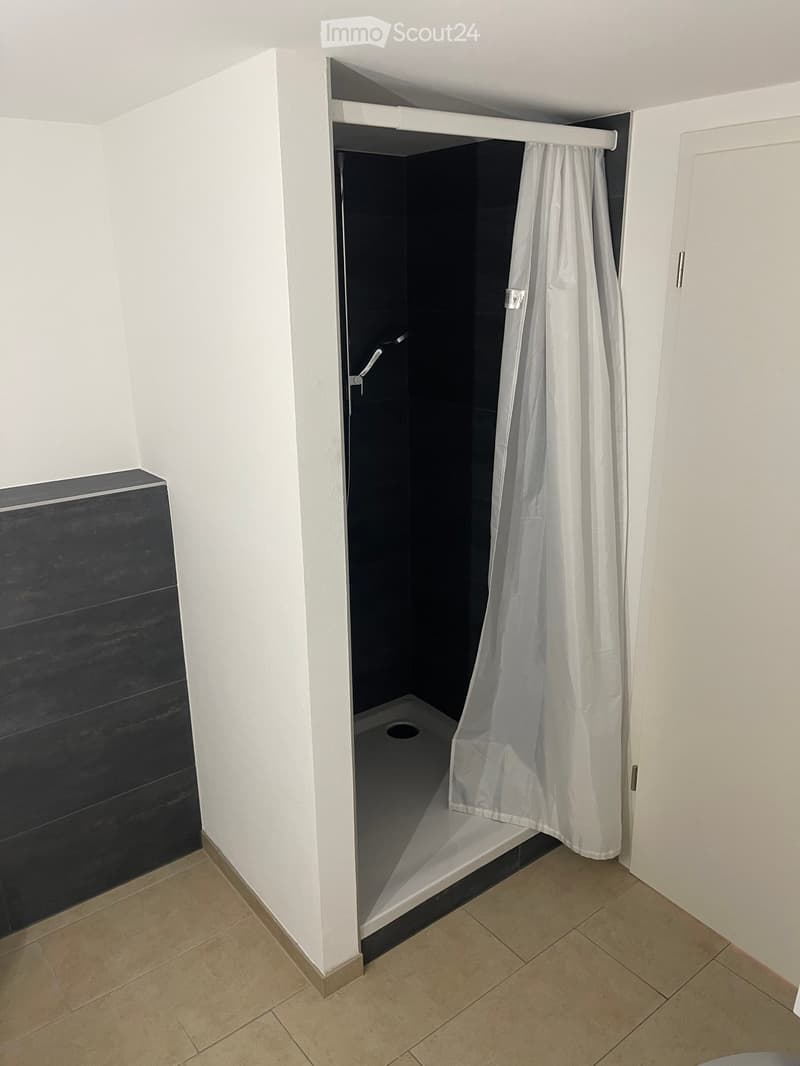 Empty bathroom with tiled floor, white walls, a shower area with a white curtain, and a dark-tiled wall section.