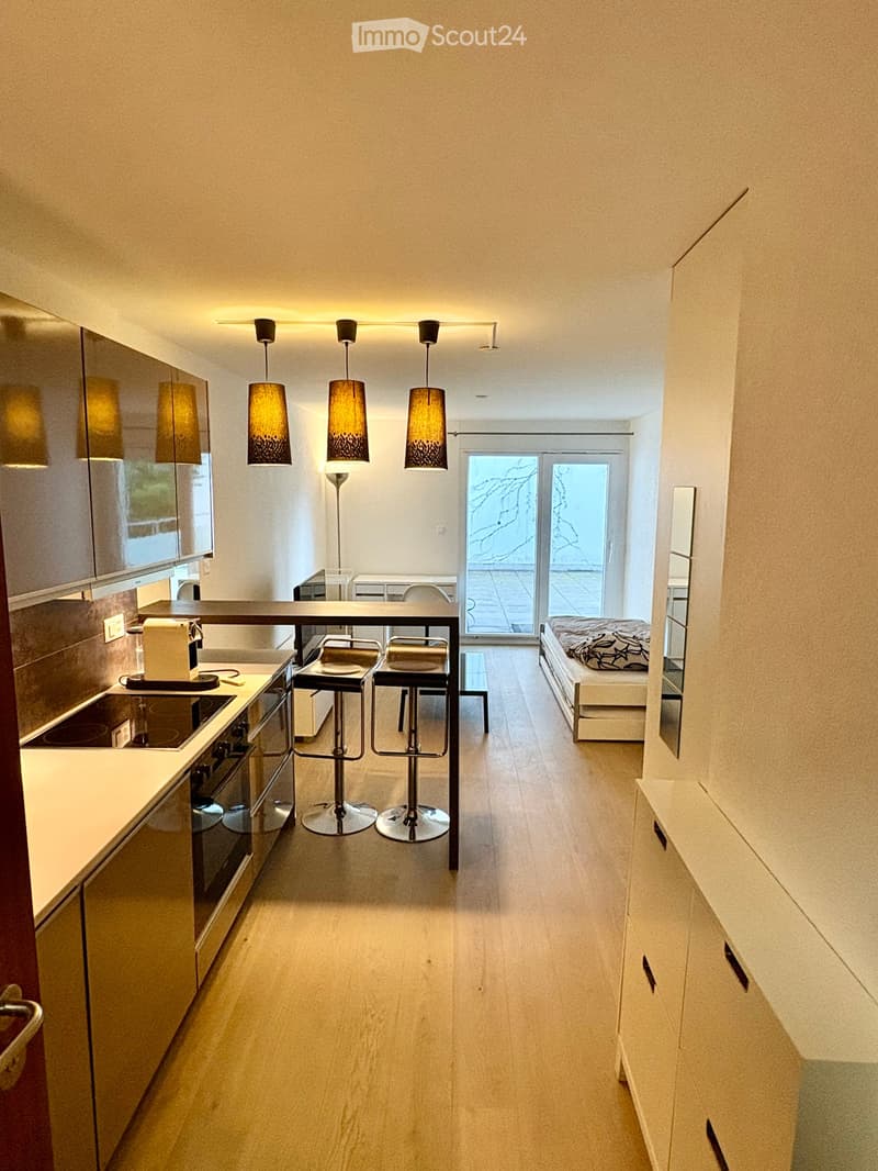 Modern open-plan kitchen and living area with light wood flooring, sleek cabinetry, and pendant lights. Features a kitchen island with stools, a bed in the adjacent space, and large windows leading to a balcony.