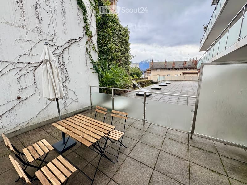 Balcony with a wooden table, four chairs, and a closed umbrella, overlooking a garden and distant mountains.