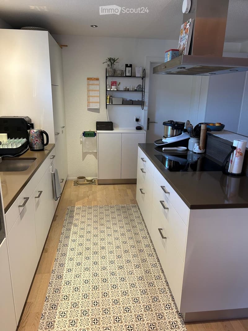 Modern kitchen with white cabinets, black countertops, and a patterned rug. Appliances include a refrigerator, oven, microwave, coffee maker, and toaster. A sink with a faucet is visible, along with a small shelf holding various kitchen items.
