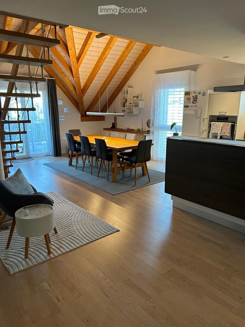 Open-plan living and dining area with wooden floors, exposed wooden beams, a dining table with chairs, a modern kitchen, and a comfortable chair with a side table.