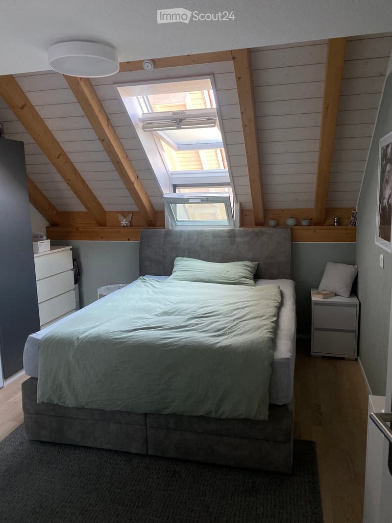Cozy attic bedroom with wooden beams, skylight windows, grey bedding, nightstands, and decorative items on shelves.