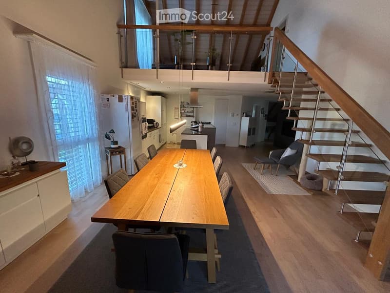 Open-plan living and dining area with a large wooden dining table, chairs, a kitchen area with a refrigerator, and a staircase leading to an upper level.