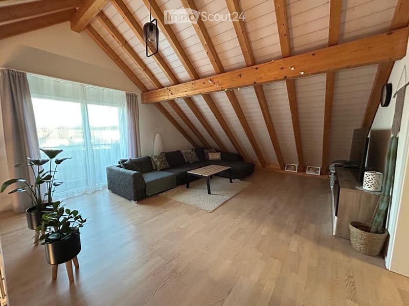 Modern living room with exposed wooden beams, large windows, grey sectional sofa, coffee table, and potted plants.